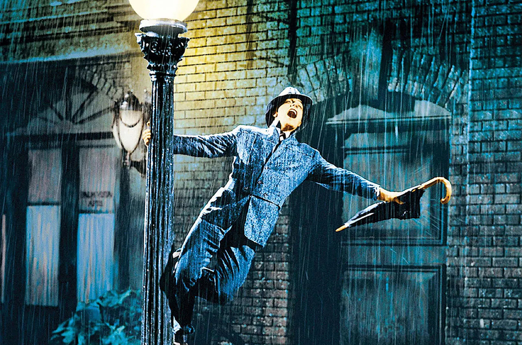 Image emblématique d’une scène de la comédie musicale « Chantons sous la pluie » représentant Don Lockwood (interprété par Gene Kelly) habillé d’un costume en tweed et coiffé d’un chapeau en feutre. Il est accroché par une main à un lampadaire de rue sous une pluie battante. Il tient un parapluie dans une main tout en souriant et chantant, exprimant une joie débordante malgré la météo. L’arrière-plan montre une rue urbaine sombre, éclairée par le lampadaire, dont les bâtiments sont en brique.