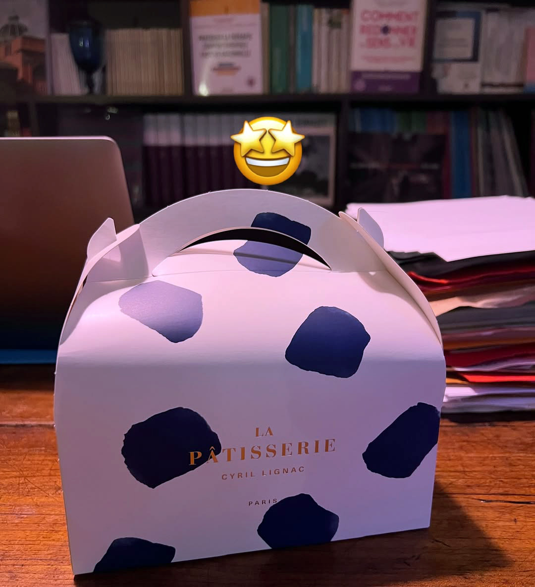 Photo d'une boîte de la Pâtisserie Cyril Lignac posée sur un bureau avec en fond une bibliothèque. L'émoticone "visage avec les yeux en forme d'étoiles" a été ajoutée sur la photo au-dessus de la boîte.