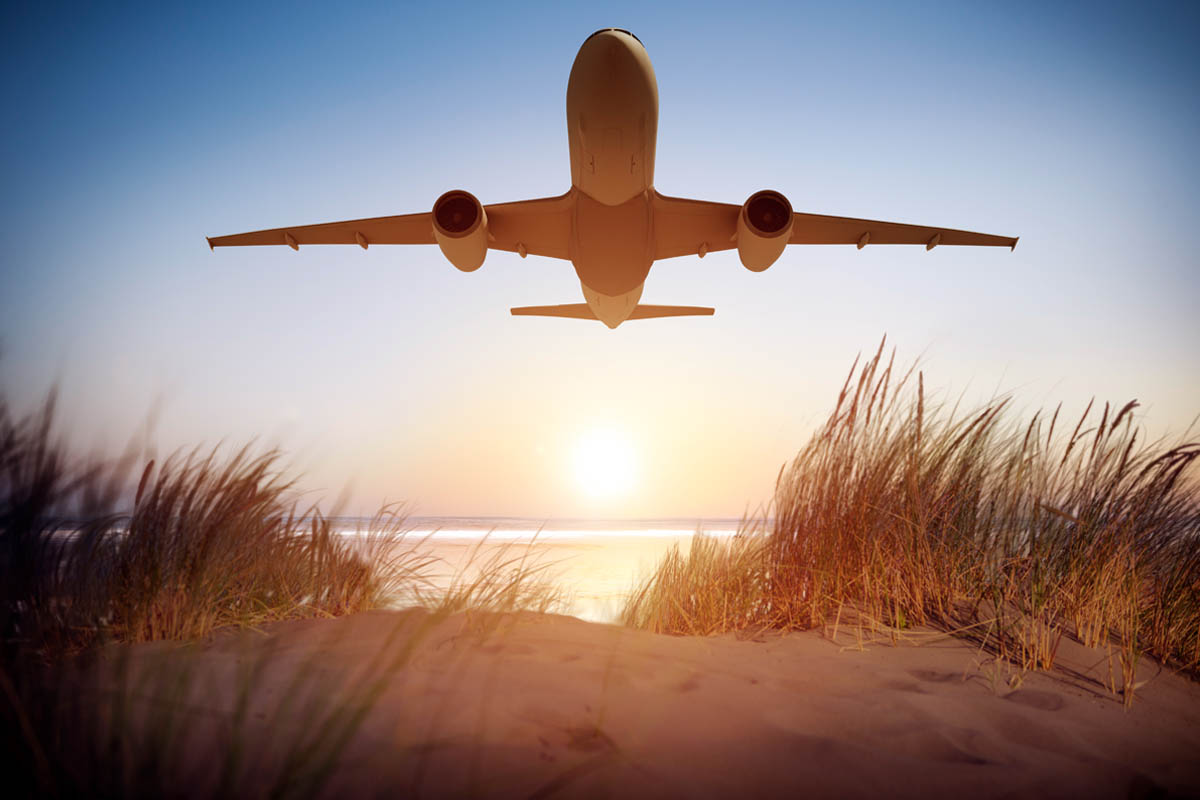 Photo d'un avion qui décolle avec au premier plan une plage de sable et de végétation, et en arrière plan, le ciel et la mer.